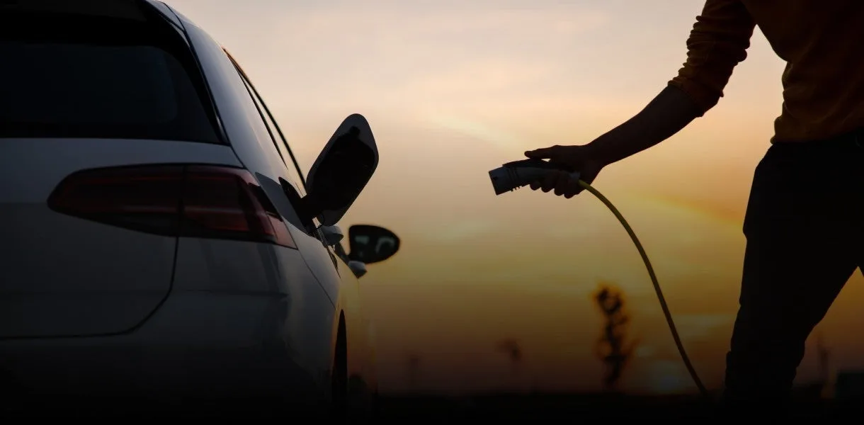 Silhouette of a driver holding a charging connector next to an EV charge port at sunset