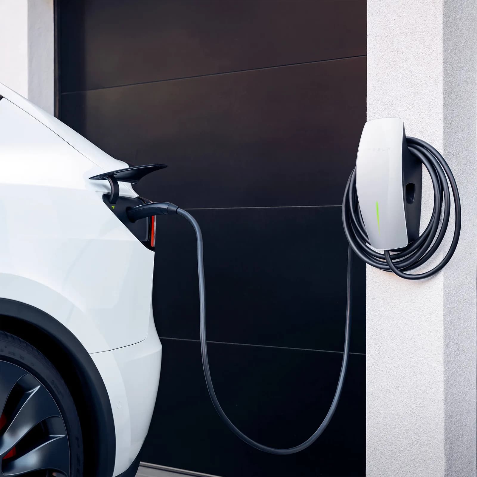 White Tesla charging from a wall-mounted Wall Connector beside a dark garage door